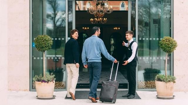 Photo of guests holding suitcase walking into hotel lobby whilst being greeted by staff member